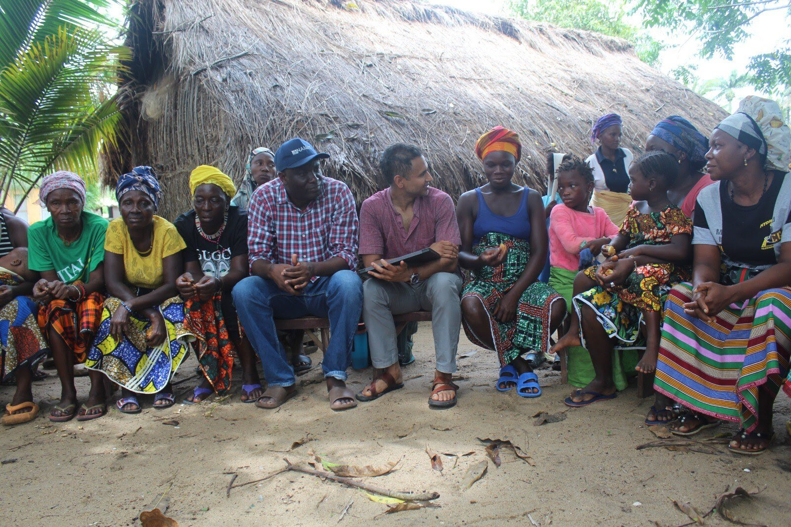 A group of people sit in a circle outside a hut.